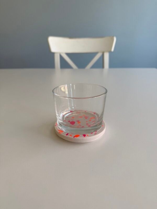 Terrazzo Glas mit weißem Deckel und pink-orangen Chips auf hellem Tisch im Studio.Elise, Stuhl und blaue Wand im Hintergrund.