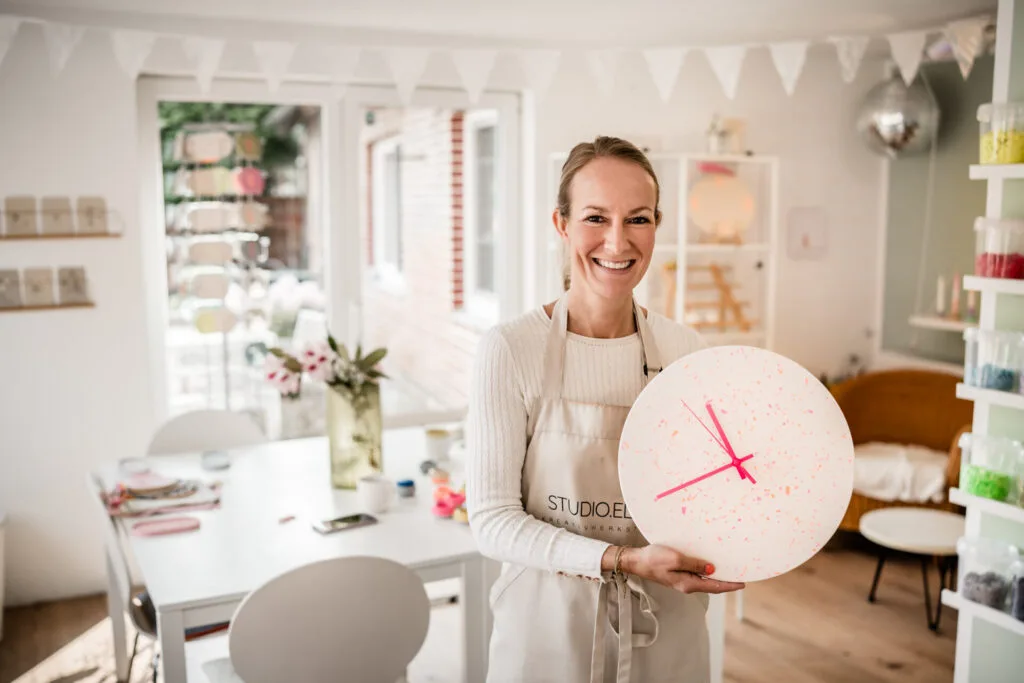 Maike im Studio Elise hält eine Terrazzo-Uhr, umgeben von Bastelmaterialien - Workshop für Terrazzo, Makramee & kreative DIY-Ideen.