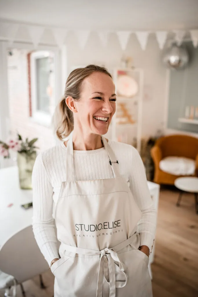 Maike im Studio.Elise, lächelnd mit STUDIOELISE KREATIVWERKSTATT-Schürze, Workshopraum für Terrazzo & Makramee, helle Deko.