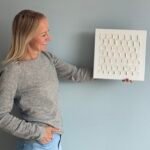 Maike im Studio.Elise hält ein weißes Texturbild aus ihrem Workshop, kreativ mit Terrazzo und Makramee, vor grauer Wand.