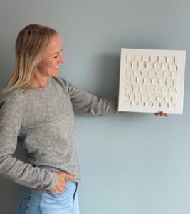 Maike im Studio.Elise hält ein weißes Texturbild aus ihrem Workshop, kreativ mit Terrazzo und Makramee, vor grauer Wand.