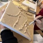 Maike gestaltet im Studio.Elise bei einem Workshop strukturierte Kunst auf Leinwand, mit Pinseln und Materialien im Hintergrund.