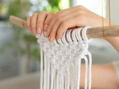 Nahaufnahme: Hände knüpfen weißen Makramee-Garn an Holzstab im Studio.Elise-Workshop bei Maike, unscharfe Pflanzen im Hintergrund.