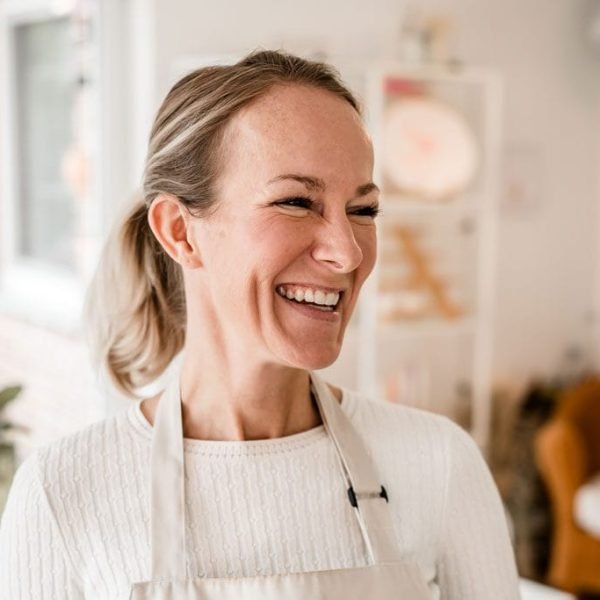 Maike im Studio.Elise, lächelnd im weißen Pullover & Schürze, bereit für Terrazzo & Makramee Workshop, gemütliches Ambiente.