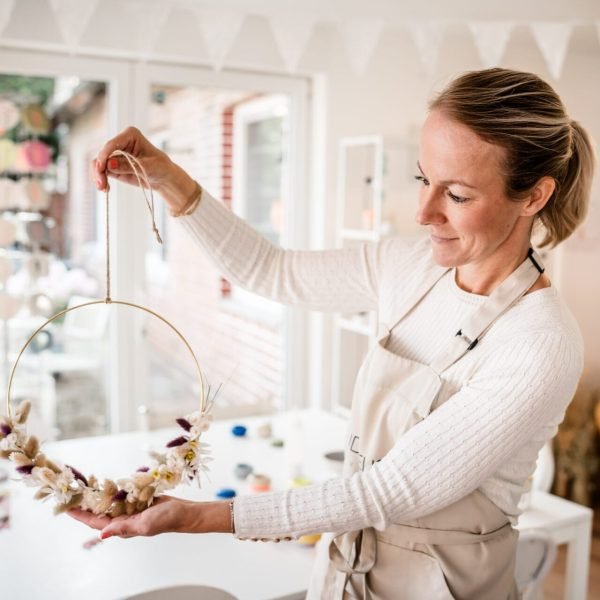 Maike im Studio.Elise hält beim Trockenblumenhoop Workshop einen getrockneten Blumenkranz, umgeben von kreativem Deko-Ambiente. Maike im Studio.Elise hält beim Trockenblumenhoop Workshop einen getrockneten Blumenkranz, umgeben von kreativem Deko-Ambiente.